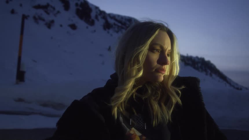 A blonde woman standing and posing outside with snowy mountains in the distance