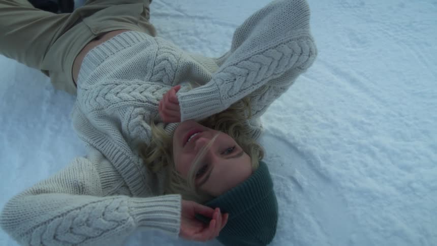 A young woman lies down on snowy ground and poses in a sweater and hat