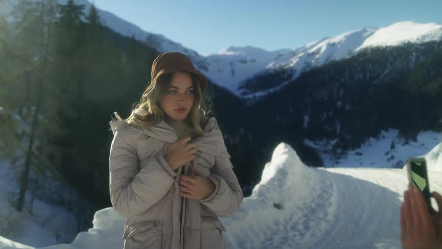 A woman with a smart phone taking a picture of a woman with a coat and hat with a mountain range sky and clouds in the background
