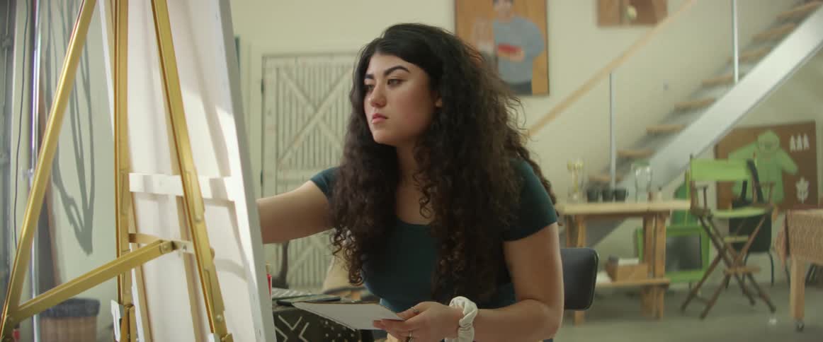 A young hispanic woman rubs her paint brush on a palette before using it to paint on a canvas held by an easel indoors during the day
