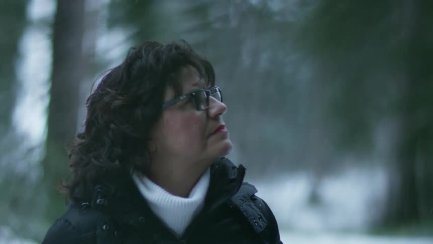 Woman with glasses and a white pullover and black coat looks up at trees in the snowy forest
