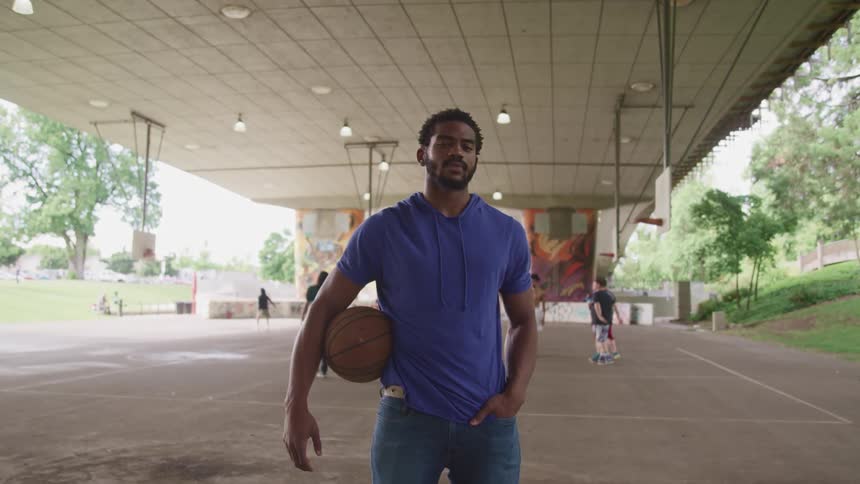 Man holding a basketball standing on a court under a bridge looking at the cmera