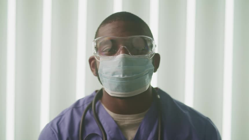 A man in blue scrubs wearing a surgical face mask and goggles standing in a hospital room looks up at the camera