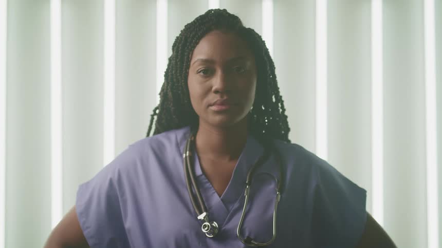 A woman with scrubs and a stethoscope in a room with a window and blinds in the background