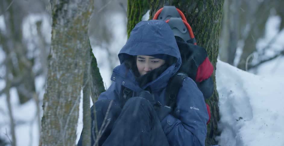 A woman with a coat and backpack sitting in a forest while it is snowing