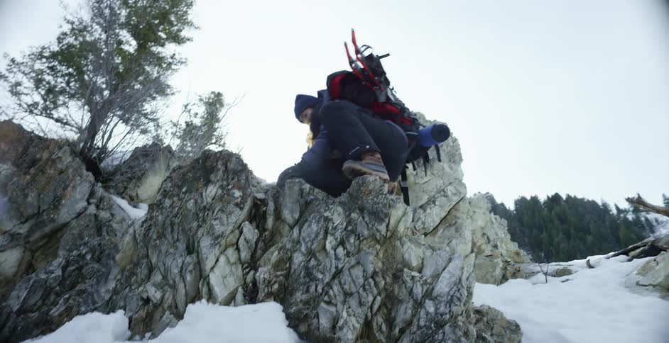 A person carrying snowshoes and a backpack climbs up and over rocks on a snowy slope