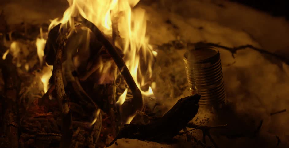 A person picks up a can of boling water using a plier next to a campfire