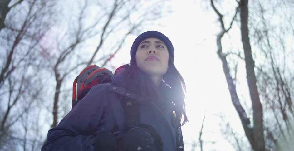 A woman with a coat hat and backpack with trees the sky and clouds in the background