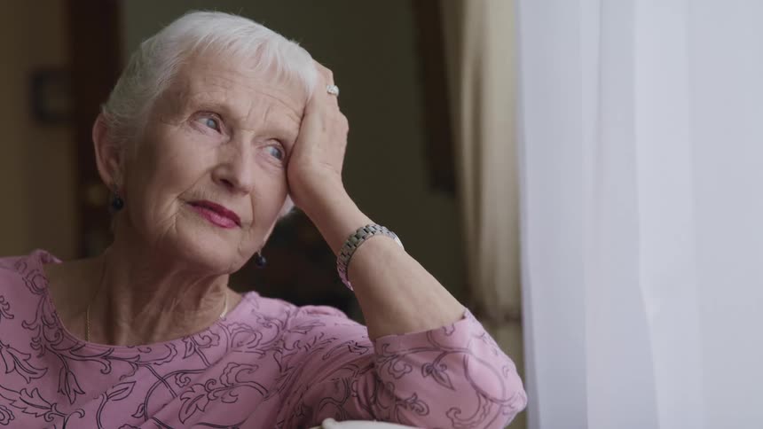 An elderly woman gazes contemplatively out of window while sitting indoors during the day