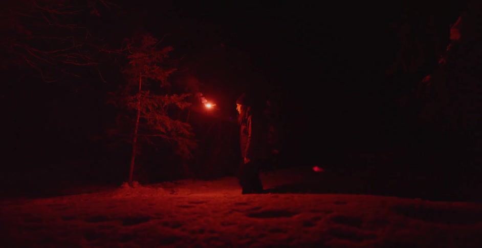 Young woman wearing a backpack lights a red flare at night close to the snow covered forest
