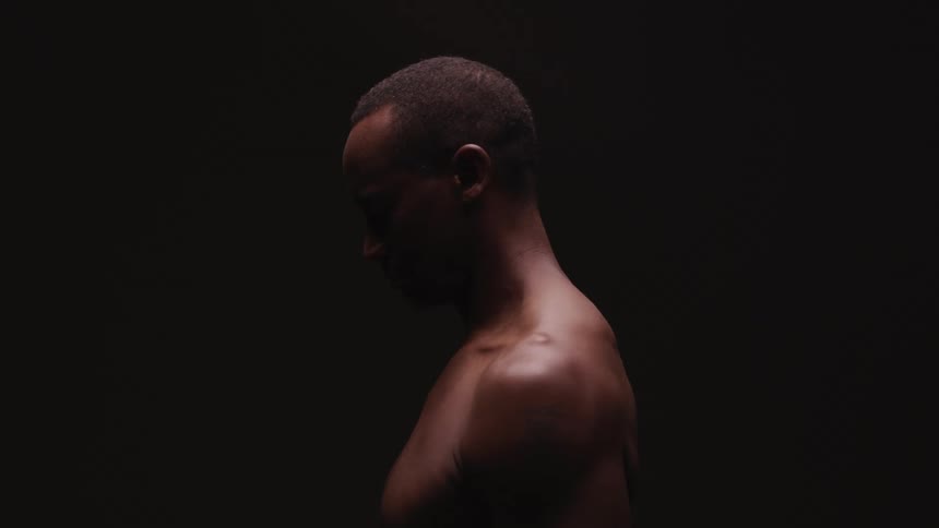A shirtless black man stands in profile and bows his head
