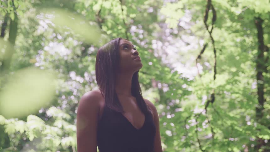 Woman with straight hair and a black shirt looks up at trees in the forest