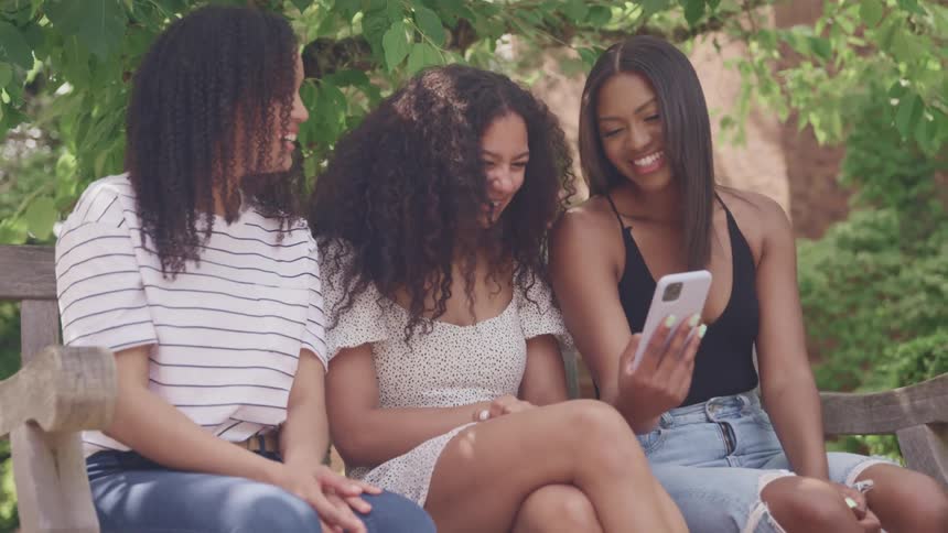 Three young women sitting on a bench outdoors at daytime laughing at a cellphone