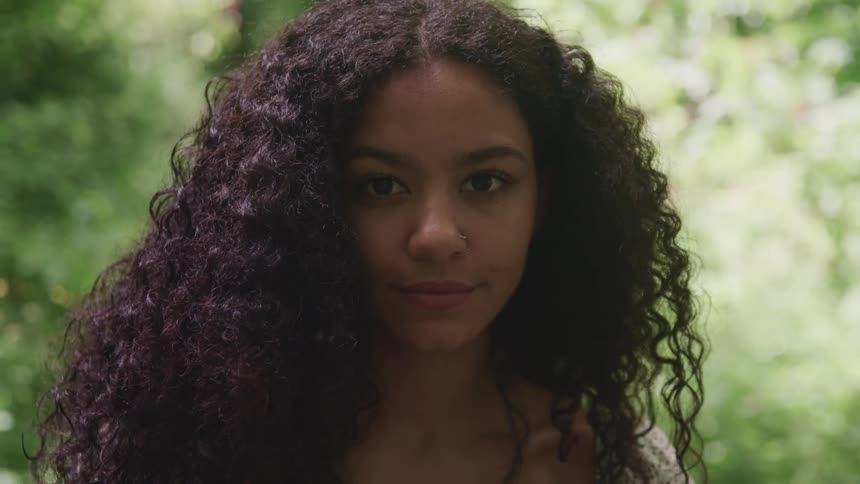 Woman with curly hair standsin woods smiling slightly