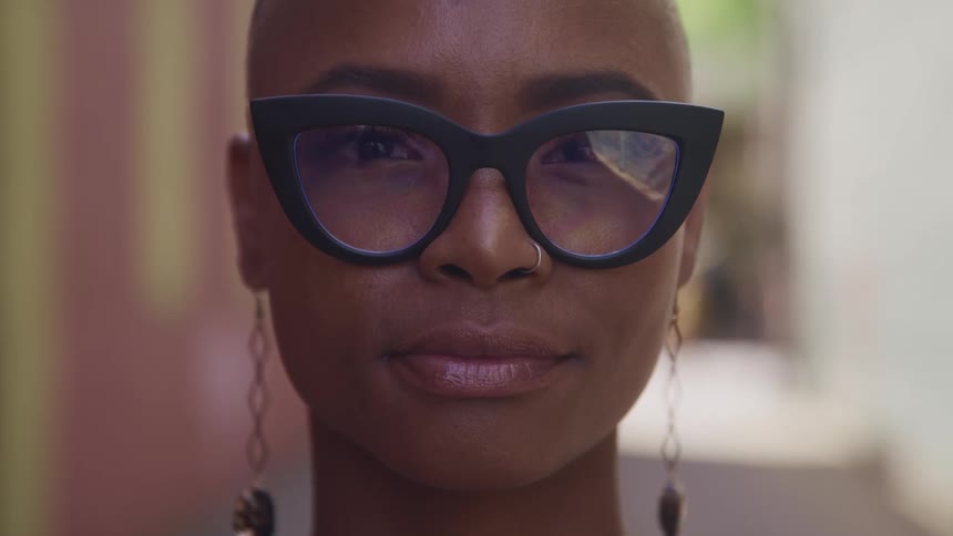 A woman with glasses a nose ring and earrings standing and staring with a alleyway in the background