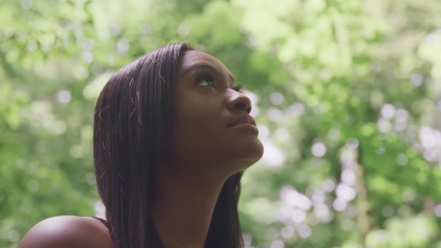 A young woman stands outdoors amidst a grove of trees while looking upwards