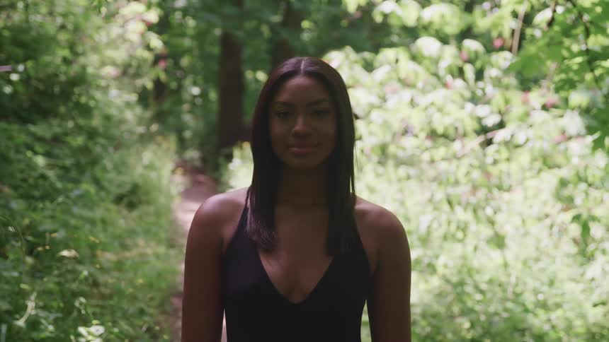 A beautiful black woman stands looking ahead with a path and trees behind her