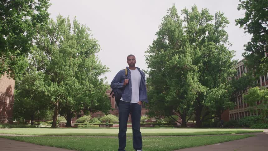 A man wearing a backpack standing outside on a lawn at a college campus at daytime looking at the camera