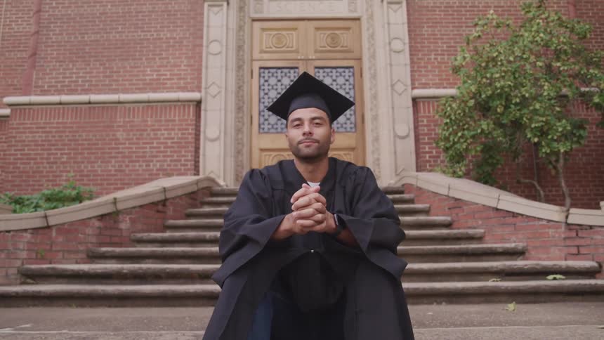A man in academic regalia turns his head to look into the camera as he sits on the stone steps in front of the door of a building