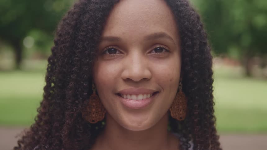 Woman wearing large carved earring stands smiling outdoors in park