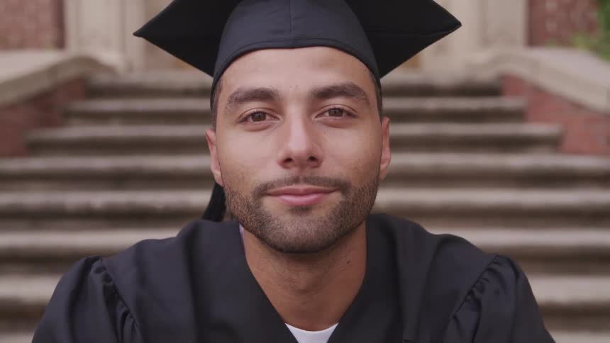 Man wearing a graduation cap and gown sitting on the stairs of a building outdoors