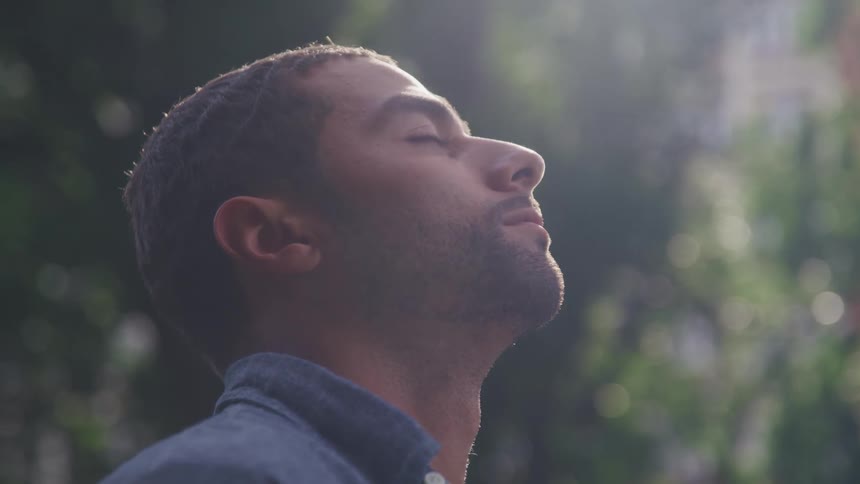 A young man looks upwards and closes his eyes while standing outdoors in the sunlight