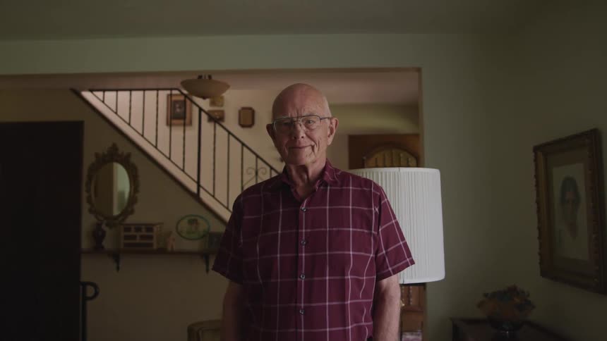 Elderly man with glasses and a red shirt faces the camera with eye contact in the living room at home