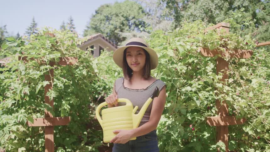 A smiling pretty young woman holding a watering can in front of a garden
