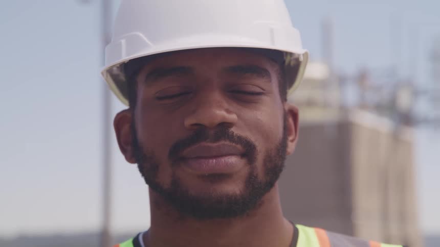 A man with a hardhat and vest with a building the sky and clouds in the background