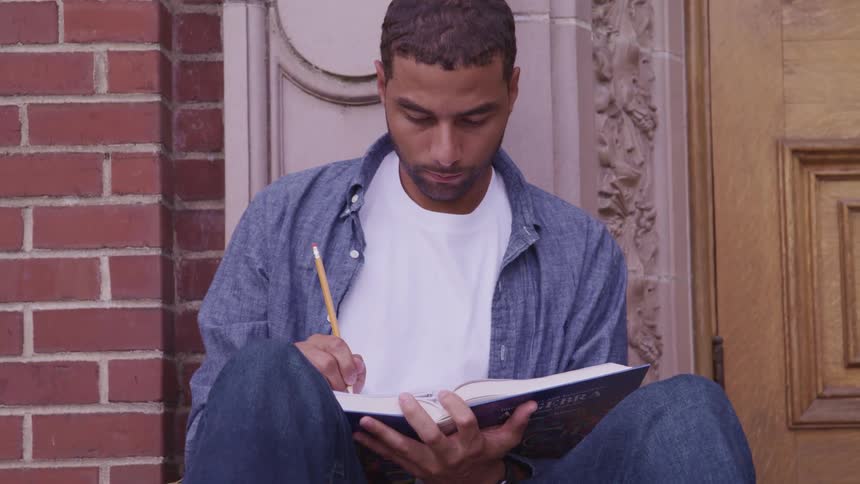 Man with a white and blue shirt writes in a notebook next to a building