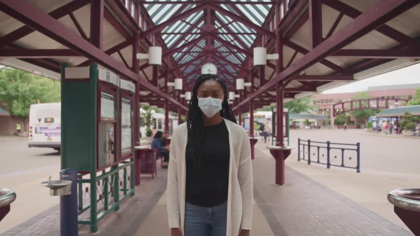 A woman stands outside under an awning at a transportation station wearing a face mask