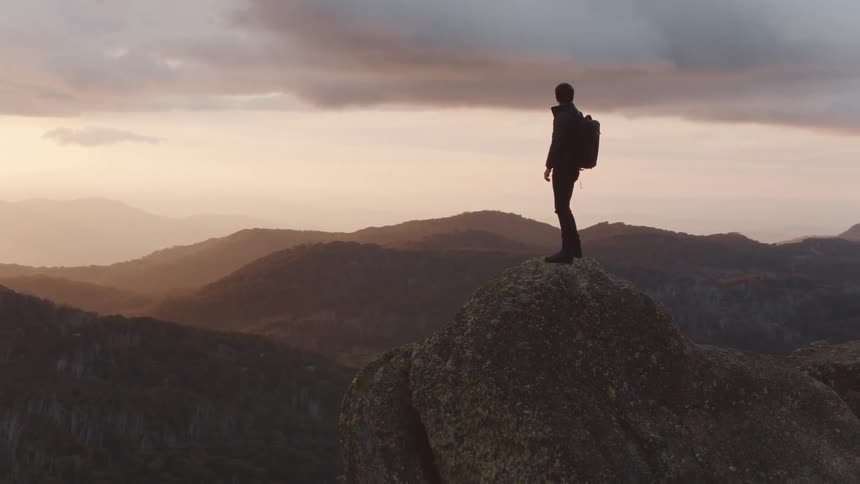 A white man with a backpack stands on a rocky peak
