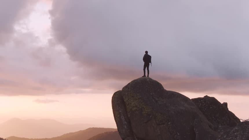 A man carrying a backpack stands high above everything on a boulder