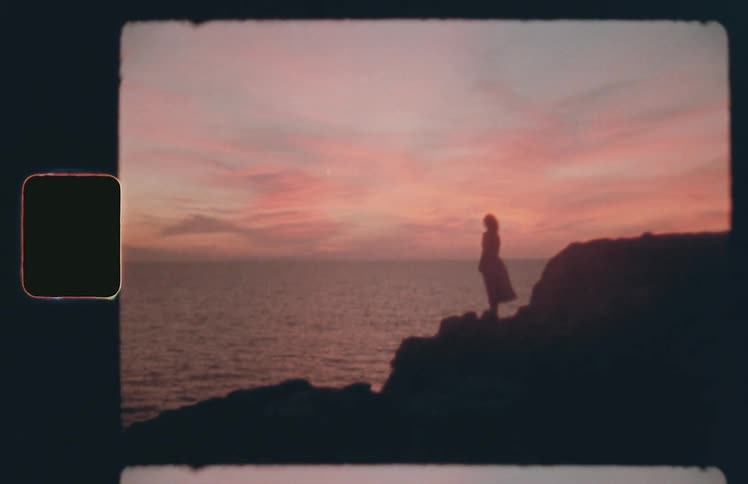 A woman in a dress stands on rocks near the coast