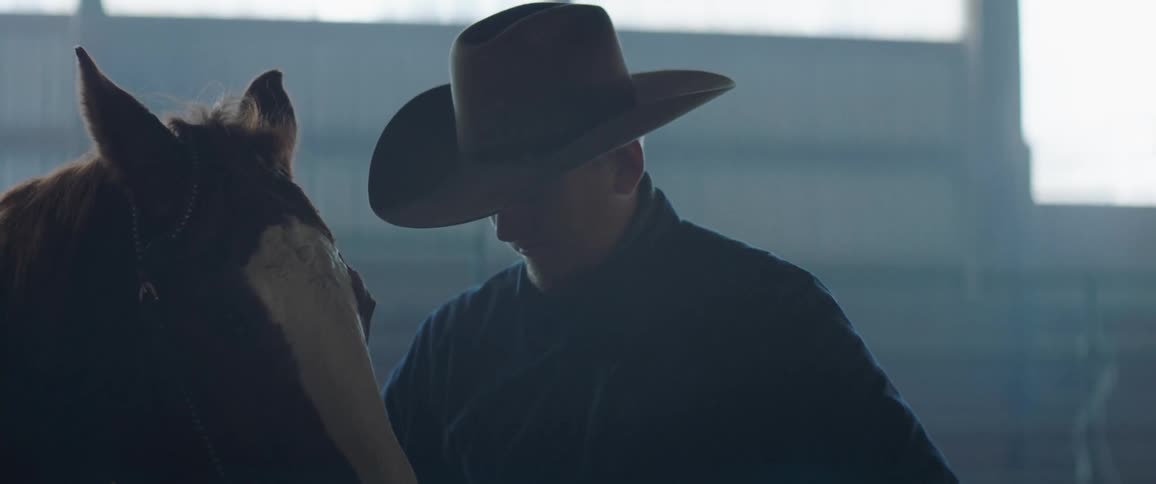 A cowboy arranging the bridle of a horse inside a stable