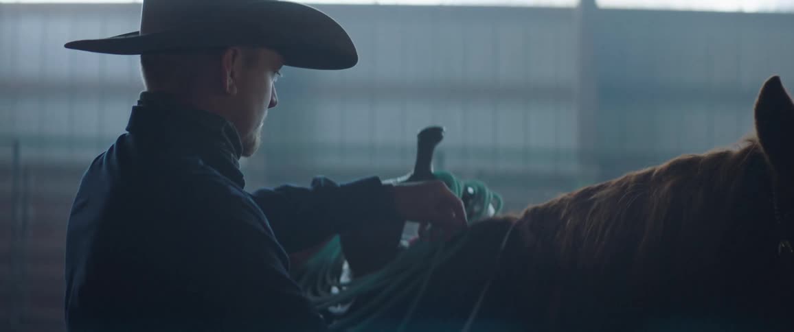A man with a hat putting a leather strap on a horse with a wall and windows in the background