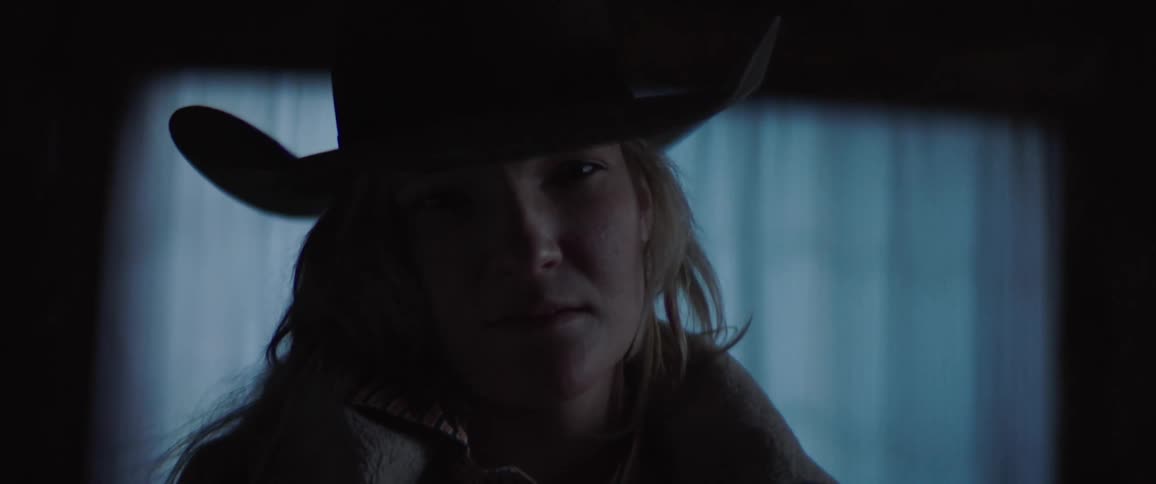 Blonde woman with a cowboy hat faces the camera with eye contact in the dark room