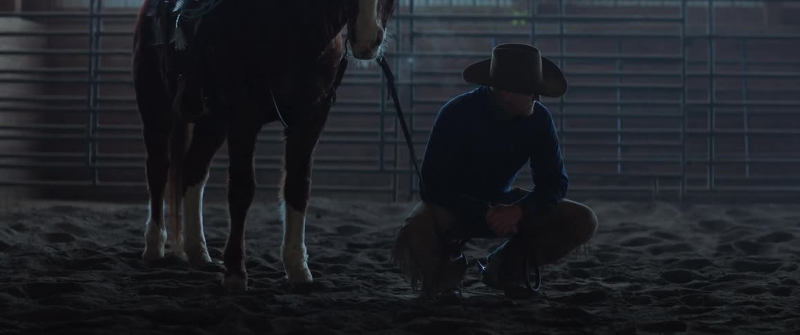Cowboy squatting on the ground next to a horse inside a stable