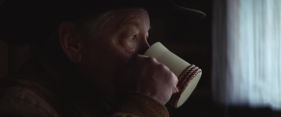 A man with a hat drinking from a mug with a wall and window in the background