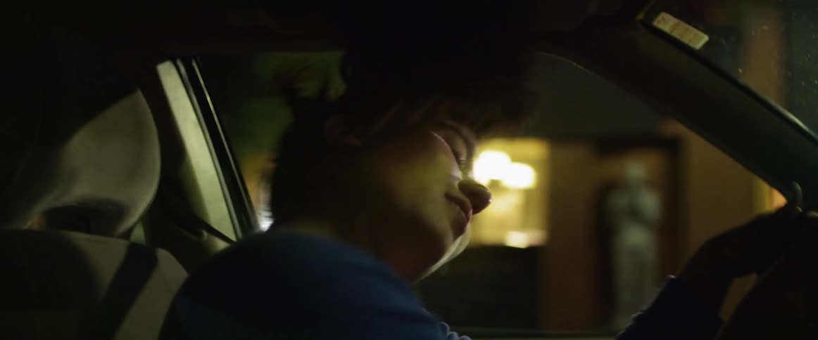 A young woman sings and whips her head around while driving through a city at night