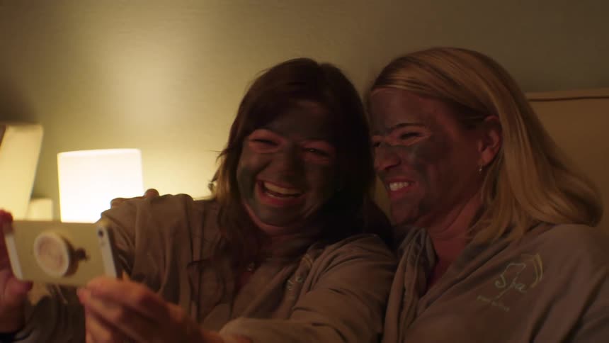 Two women smiling and laughing while wearing mud masks in a spa and posing for a selfie