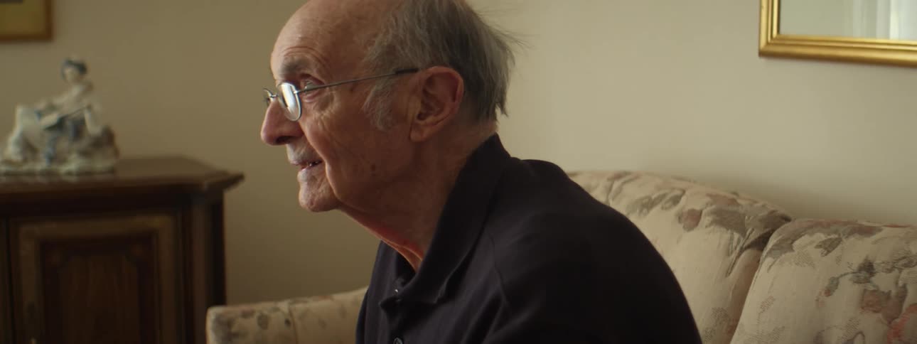 Old bald man with glasses and a black polo shirt watches tv in the living room at home