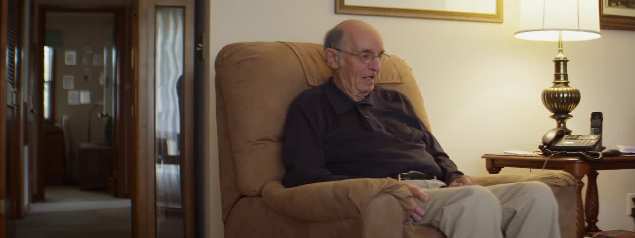 A man with glasses sitting on a couch with a wall and doorway in the background
