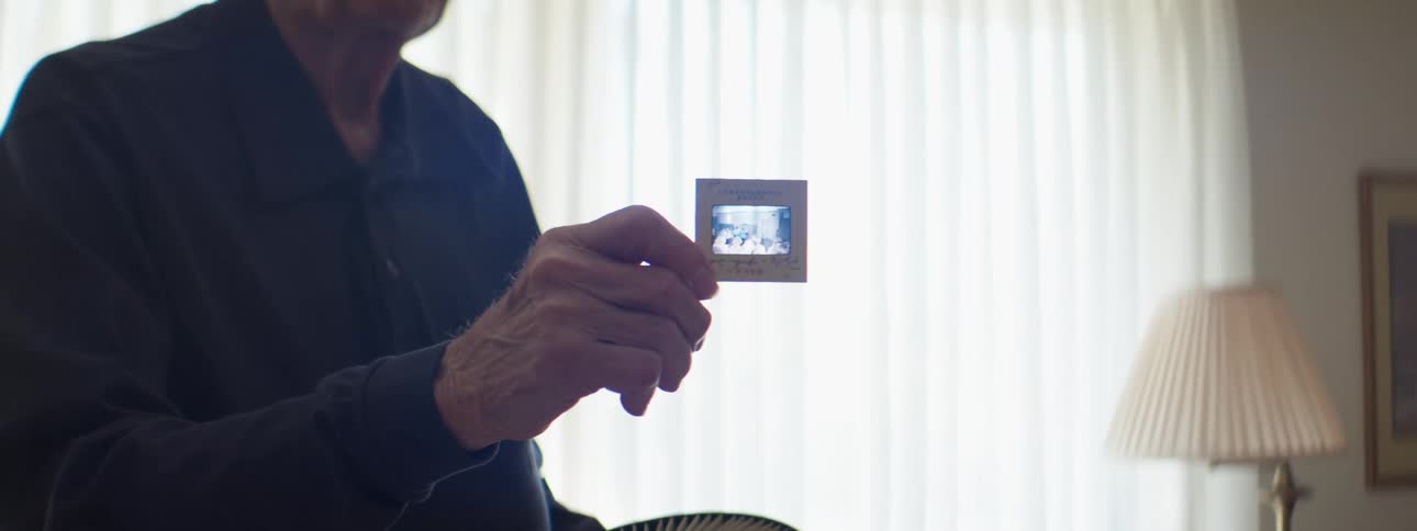A man in a room holding a projector slide with a lamp and window in the background