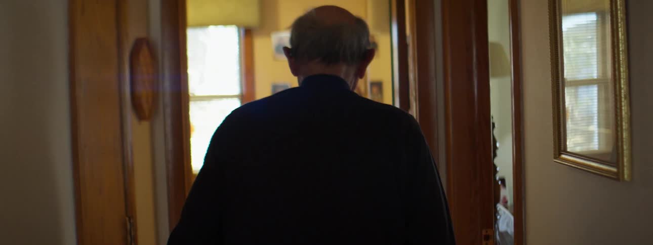 A man walks down the hallway of his house lined with picture frames and into an office with more picture frames of children