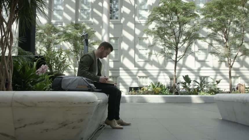 A man sits and reads on a marble bench in an outdoor atrium filled with plants