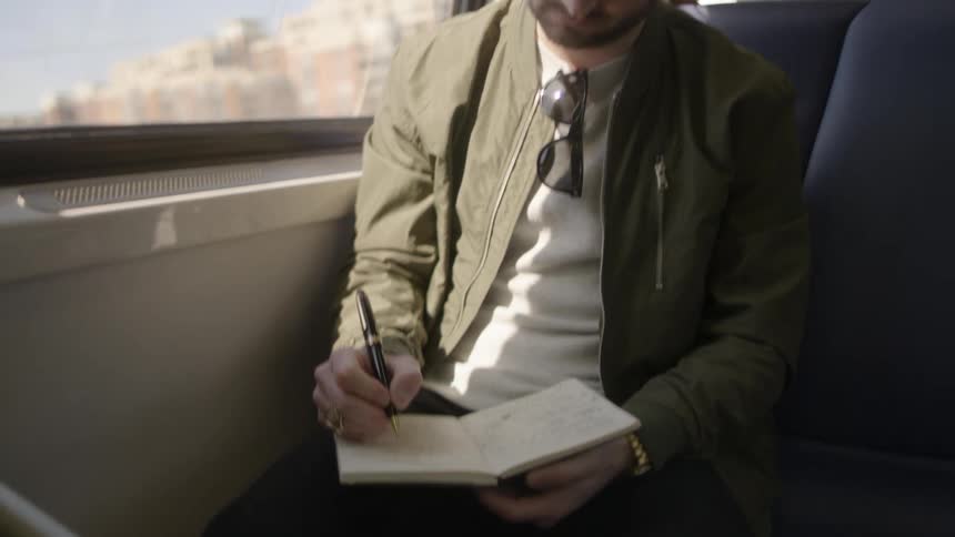 A man writes in a notebook while riding in a train during the day