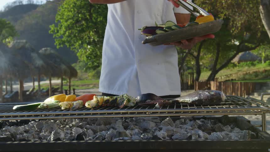 A chef places vegetables and seafood on an open grill on the beach