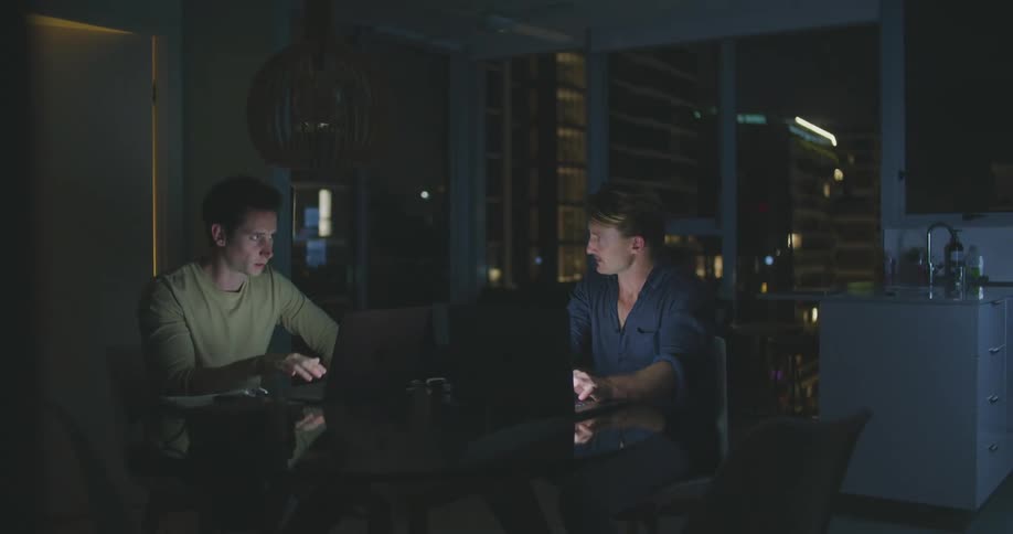 Two men are sitting in a room and on laptops