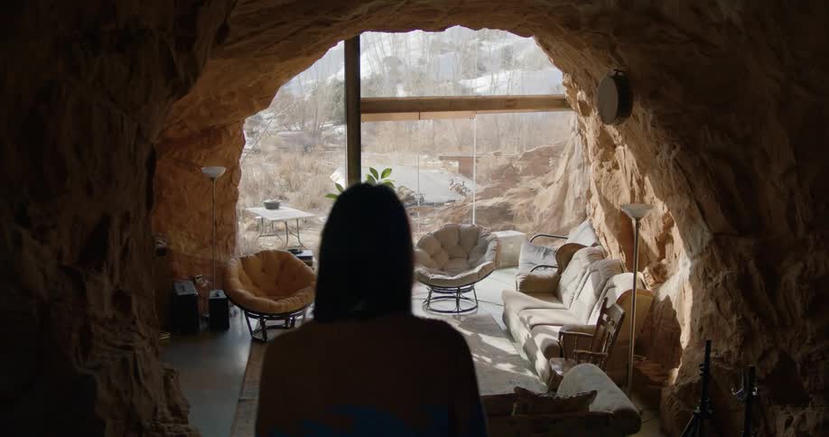 A woman walks down the steps to a living room made of rock walls and glass
